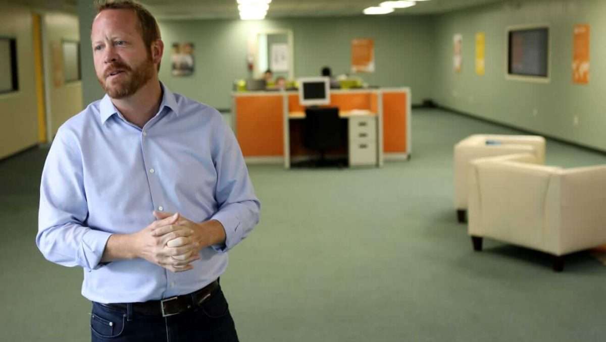 A man stands speaking, hands clasped, in an open office space with cubicles, chairs, and soft lighting in the background.