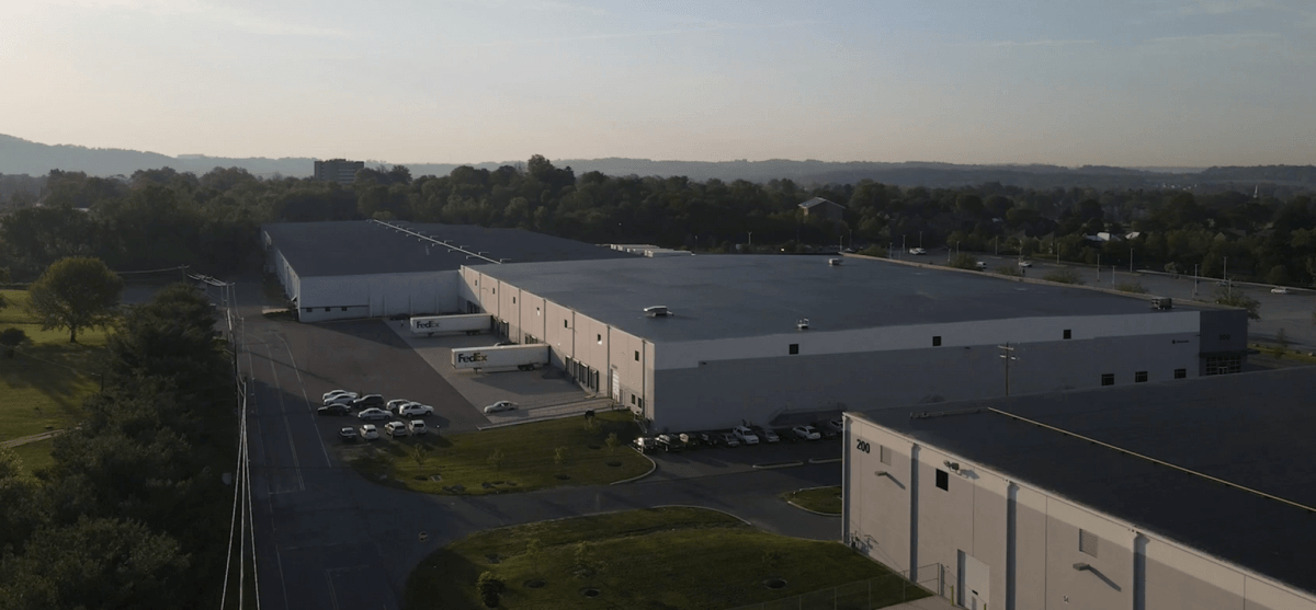 A large warehouse building framed by parked FedEx trucks; surrounded by numerous cars in an industrial area with distant hills and trees under a morning sky.