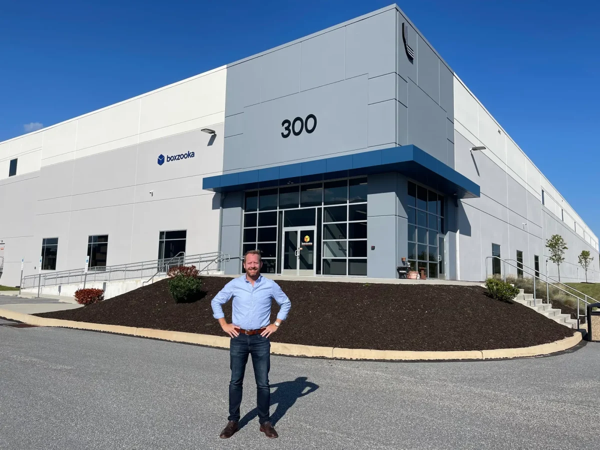 A man stands confidently outside a large, modern industrial building labeled "boxzooka" with the number "300" on the facade, under a clear blue sky.