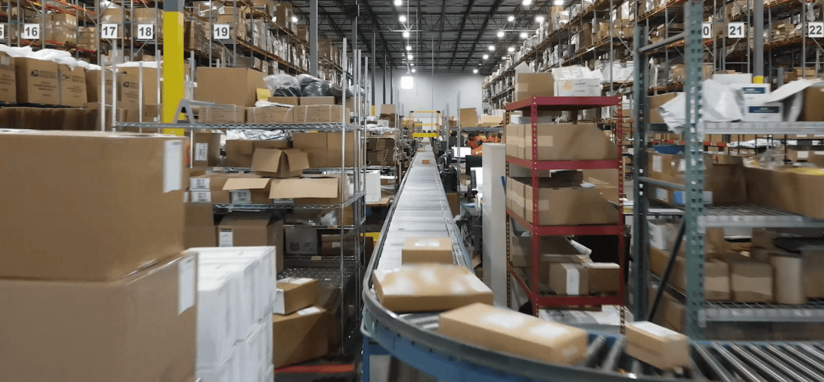 Conveyor belt transports boxes through a busy warehouse filled with shelves stacked with various packages, under a high ceiling with bright overhead lights. Numbered aisles for organization are visible.