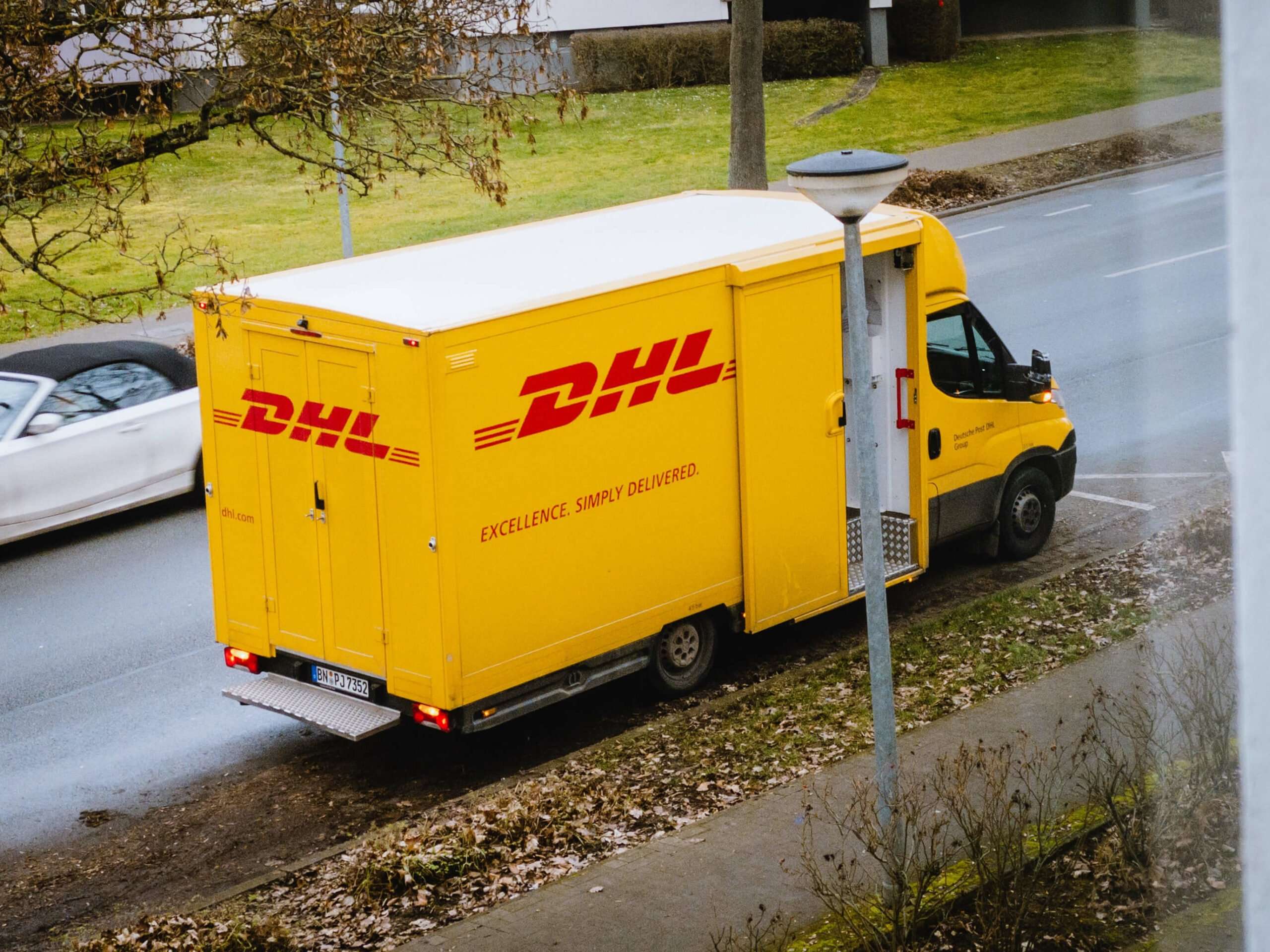 Yellow DHL delivery van parked on a roadside curb; logos and "EXCELLENCE. SIMPLY DELIVERED." text visible; adjacent gray road with passing white car; overcast suburban setting.