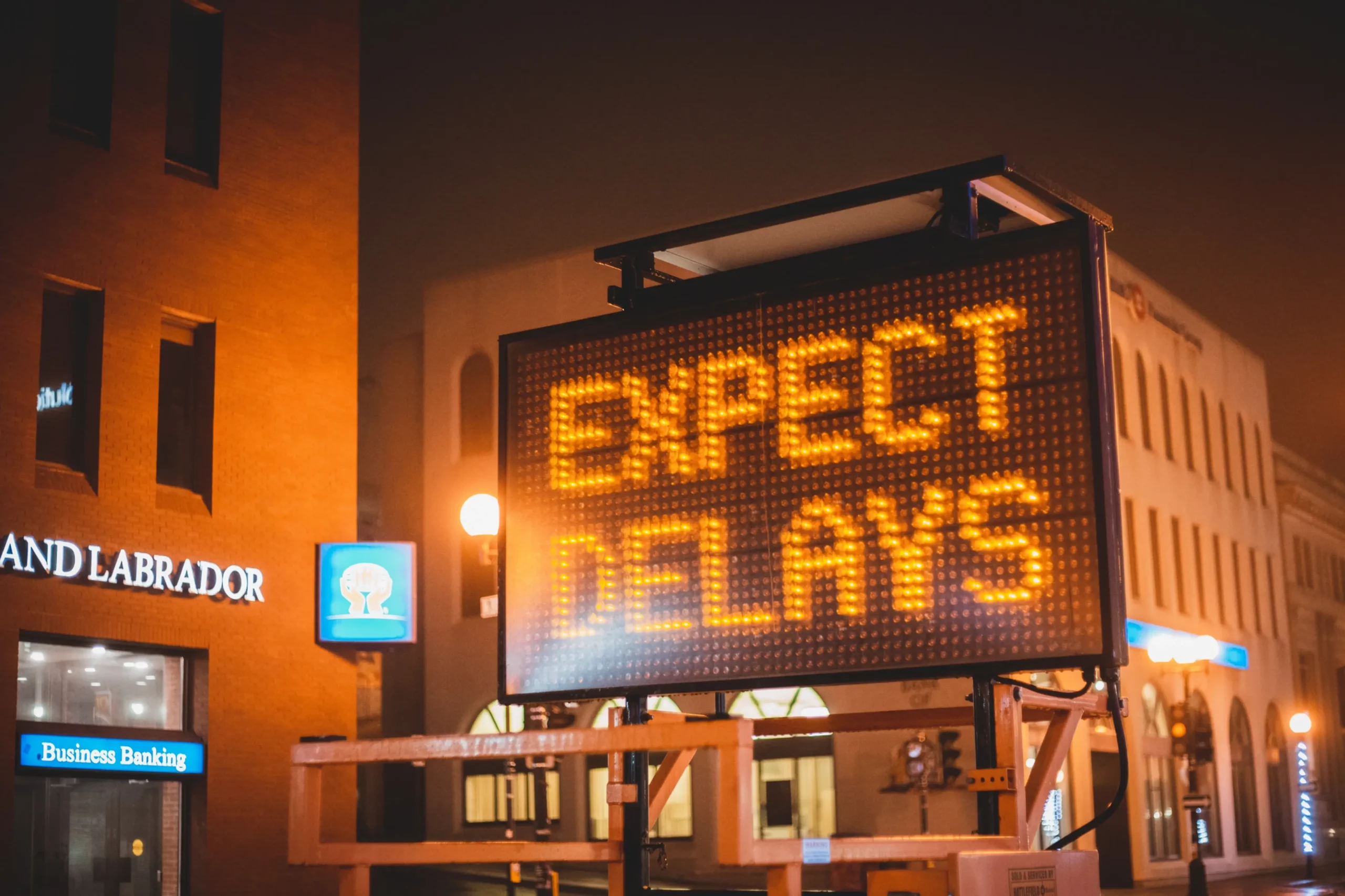 Electronic sign displaying "EXPECT DELAYS" glows in a city street at night, surrounded by buildings and illuminated by streetlights.