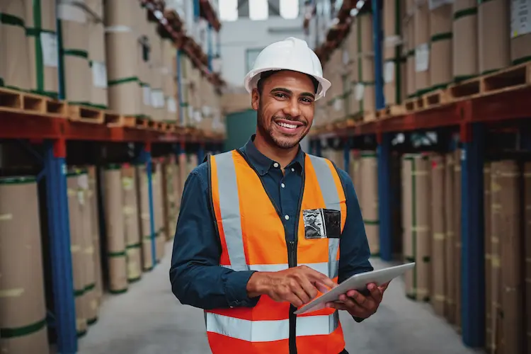 Smiling warehouse supervisor holding a tablet between rows of stored inventory