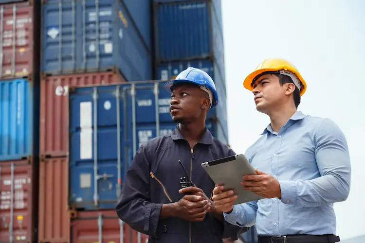 Two logistics workers with a tablet in front of stacked shipping containers