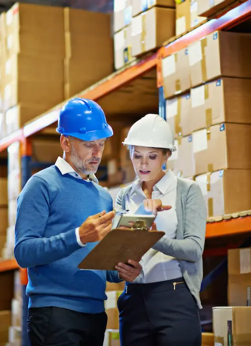 Two warehouse managers reviewing a clipboard and tablet in front of pallet racking
