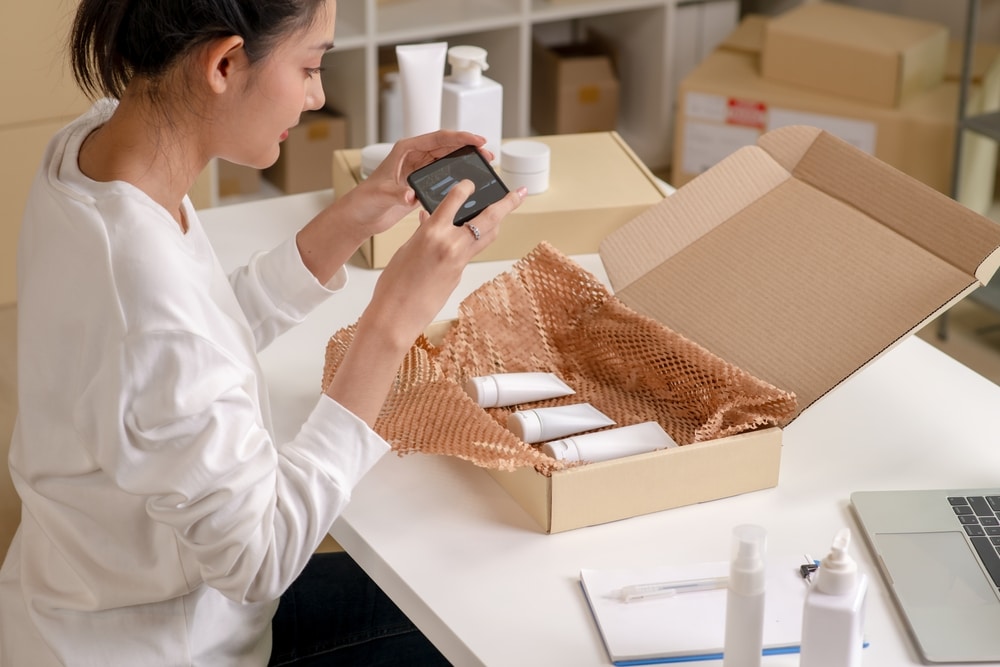 Asian e-commerce seller taking photo of the prepared orders with smartphone and laptop in a warehouse setting for an online beauty shop